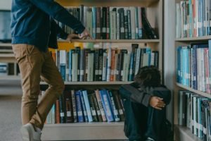 a photo of a person sitting on the corner beside a wooden bookshelf with another person pointing at them